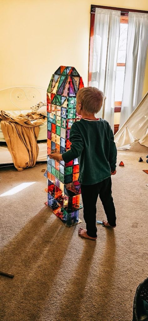 A young boy standing barefoot in a bedroom, admiring a tall tower toy he built with colorful tiles.