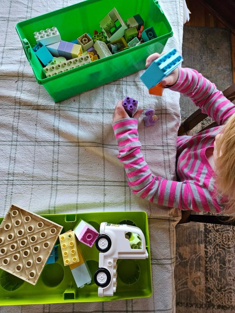 Child in a pink striped shirt playing with large plastic building blocks at a table with a green container full of toy bricks.