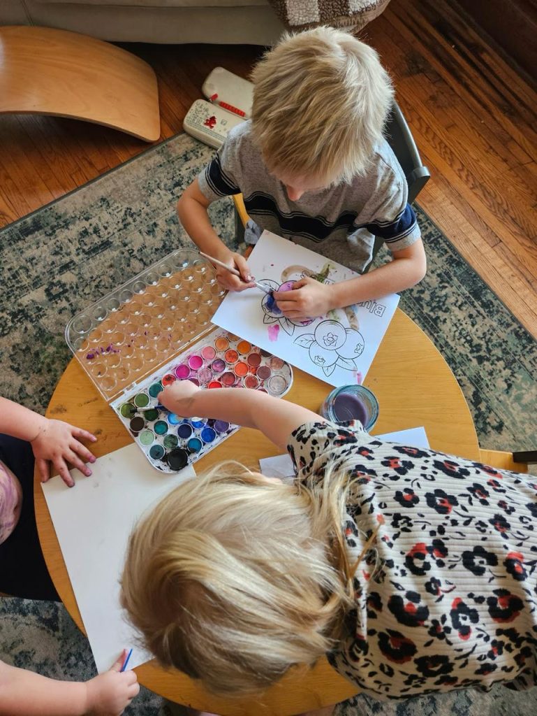 Image of three young children color painting a drawing in a paper on top of round wooden table.