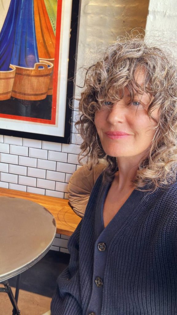 Ninka wearing a dark cardigan, sitting indoors at a café with a colorful painting in the background.
