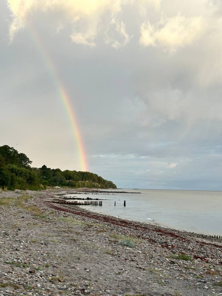 A calm shoreline with pebbles, greenery on the left, and a rainbow stretching down toward the horizon.